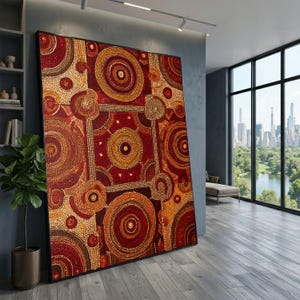 Puede incluir: Gran obra de arte enmarcada con un patrón geométrico en tonos rojos, naranjas y dorados. El diseño presenta círculos concéntricos y formas cuadradas, creando una apariencia texturizada. La obra de arte está apoyada contra una pared gris en una habitación moderna.