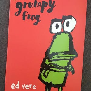 以下が含まれることがあります： エド・ヴェール著の子供向け絵本「Grumpy Frog」で、鮮やかな赤色の表紙です。イラストには、大きな白い目と黒い輪郭を持つ、様式化された緑色のカエルが描かれています。表紙の左上隅には小さなパフィンのロゴが見えます。