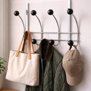 May include: A chrome over-the-door coat rack with six black ball-shaped hooks. A beige tote bag with brown leather handles, a green quilted jacket, and a tan baseball cap hang from the hooks. The rack is mounted on a light-colored wall.