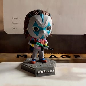 May include: A stylized vinyl figure wearing a white mask with blue diamond patterns, brown hair, a pink and blue patterned shirt, and grey trousers. The figure holds a green rifle and stands on a grey hexagonal base with a plaque labeled Nickname.