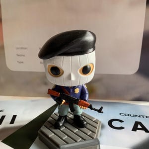 Puede incluir: Una figura de vinilo estilizada que lleva una boina negra y una máscara blanca con cuencas oculares negras. El personaje viste un uniforme azul con una insignia dorada, pantalones verde claro y botas negras, sosteniendo un rifle negro y marrón.