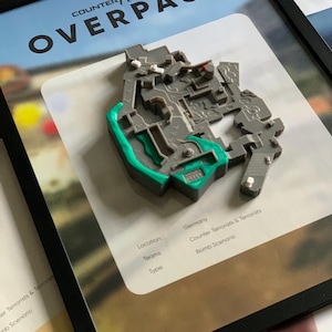 May include: Framed artwork featuring a 3D model of the Overpass map from Counter-Strike. The map is gray with green accents, set against a white background with the game's logo and text. The frame is black.