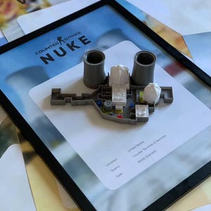 May include: A framed display featuring a 3D model of the Counter-Strike Nuke map. The model includes two grey cooling towers, white reactor buildings, and grey structural pathways. The background print displays the text Counter-Strike Nuke.