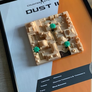 May include: A framed 3D map of the Counter-Strike Dust II game level in beige, featuring small green palm trees. The print includes the text Counter-Strike Dust II, Location: Morocco, Teams: Counter Terrorists & Terrorists, and Type: Bomb Scenario.
