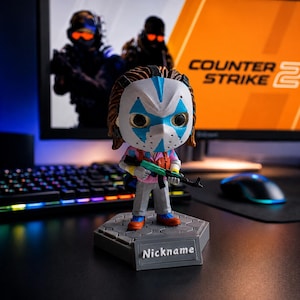 May include: A stylised vinyl figure wearing a white mask with blue accents, a pink shirt, and grey trousers, holding a small green and black rifle. The figure stands on a hexagonal grey base with the word Nickname printed on a raised rectangular plaque.