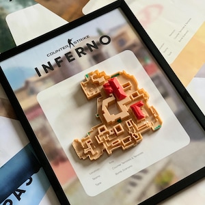 Puede incluir: Una pieza de arte mural 3D enmarcada que presenta el diseño del mapa Inferno de Counter-Strike. El modelo de plástico beige incluye detalles de techos rojos sobre un fondo blanco con texto negro que detalla la ubicación en Italia, equipos y escenario.