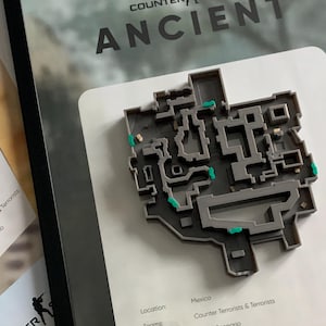 Puede incluir: Una pieza de arte mural 3D enmarcada que presenta el diseño del mapa Ancient de Counter-Strike. El mapa de plástico gris incluye pequeños acentos verdes y está montado sobre un fondo blanco con el texto: Location: Mexico, Teams: Counter Terrorists & Terrorists, Type: Bomb Scenario.