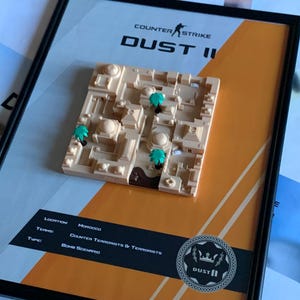 May include: A framed 3D wall art piece featuring a beige miniature map of the Counter-Strike Dust II game level. The display includes small green palm trees, a brown area, and text reading Counter-Strike DUST II, Location: Morocco, Teams: Counter Terrorists & Terrorists, Type: Bomb Scenario.