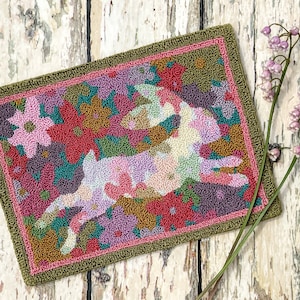 Puede incluir: Una alfombra de crochet colorida con un conejo rosa estilizado saltando a través de un campo de flores. Las flores son en tonos de rosa, rojo, verde y marrón.