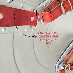 May include: Close-up of a section of metal armor with red leather straps and rivets. The text "COMFORTABLE LEATHER GRIP FOR EASE OF USE" is visible. The armor has a silver finish and is designed for protection.