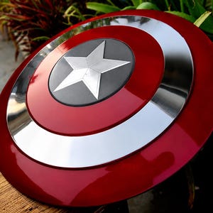May include: A red, white, and silver shield with a star emblem. The shield has a glossy finish and a circular design. The star is silver and set against a black background. The shield is a prop or collectible.