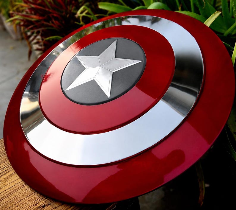 May include: A red, white, and silver shield with a star emblem. The shield has a glossy finish and a circular design. The star is silver and set against a black background. The shield is a prop or collectible.