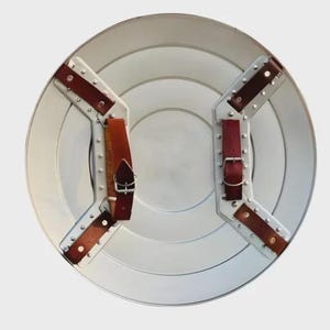 May include: A silver-colored, round shield with concentric circles and riveted metal supports. It features two sets of brown leather straps with silver buckles for securing the shield. The shield has a classic, vintage aesthetic.