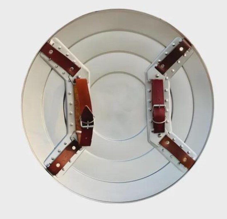 May include: A silver-colored, round shield with concentric circles and riveted metal supports. It features two sets of brown leather straps with silver buckles for securing the shield. The shield has a classic, vintage aesthetic.