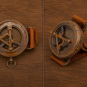 May include: A brass sundial compass featuring a circular face with Roman numerals and a central gnomon, attached to a tan leather strap. The device includes a small metal ring at the base and a hinged mechanism, displaying a weathered metallic finish.