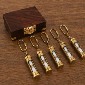 May include: Five brass hourglass keychains with white sand, each featuring a cylindrical metal casing and a secure lobster clasp attachment. A dark brown wooden box with a gold-toned metal latch is positioned behind the keychains.