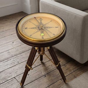 May include: A round side table featuring a tabletop designed as a large nautical compass with a gold-toned rim and a central needle. The table is supported by three dark brown wooden legs connected by brass-colored metal hardware and struts.