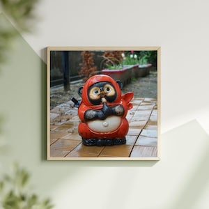 May include: A ceramic ninja Tanuki statue, featuring a red hooded outfit, black gloves, and a small sword. The statue has a white belly and is set against a blurred outdoor background. The statue is a traditional Japanese folk figure.
