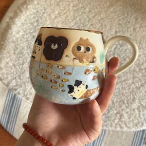 Könnte beinhalten: Handbemalte Keramik-Tasse mit einem skurrilen Design. Die Tasse hat einen hellblauen Boden mit Fisch- und Tierillustrationen. Der obere Rand ist hellbraun und der Griff ist weiß. Die Tasse wird von einer Person gehalten.