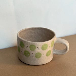May include: A beige ceramic mug with a rounded handle. The mug is decorated with green polka dots and small cross-like designs. The mug is suitable for coffee, tea, or other beverages.