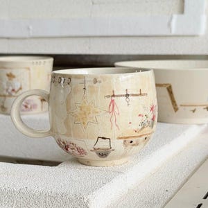 May include: Cream-colored ceramic mug with a handle, featuring hand-painted illustrations of a kitchen scene with a star, a pot, and a hanging rack. The mug has a rounded shape and a light beige background.