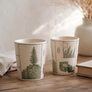 Könnte beinhalten: Zwei cremefarbene Keramikbecher mit handgezeichneten Illustrationen. Ein Becher zeigt einen Baum auf einem grasbewachsenen Hügel, der andere eine Fensterszene mit einem Stuhl. Die Becher sind zylindrisch und stehen auf einer Holzoberfläche.