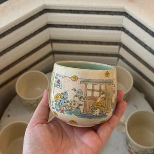 Könnte beinhalten: Eine handbemalte Keramiktasse mit einer skurrilen Illustration eines Hauses mit Blumen und einem blau-grünen Innenraum. Die Tasse hat einen hellbeigen Hintergrund mit einem hellgrünen Innenraum.