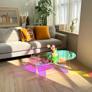 May include: A modern living room scene featuring a gray sofa with orange and yellow throw pillows. A colorful, oval-shaped coffee table reflects light, creating a rainbow effect. A framed artwork and plants add to the decor.