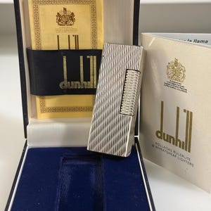 May include: A silver-toned Dunhill Rollagas lighter with a textured wavy pattern, presented in a dark blue display box. The set includes a yellow instructional card with the Dunhill logo and a white booklet titled Dunhill Rollagas Rulerlite.