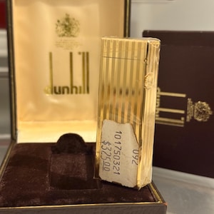 May include: A gold Dunhill lighter with vertical stripes, presented in its original box. The box is brown with a gold interior and the Dunhill logo. A price tag is attached to the lighter.