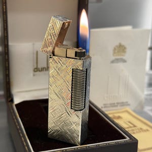 May include: A silver lighter with a textured pattern, lit with a blue and yellow flame. The lighter is in an open brown box with a dark interior. The brand name "dunhill" is visible on the box.