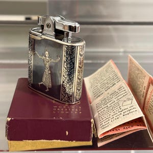 May include: Vintage Ronson lighter with a silver finish and a decorative panel featuring a detailed illustration of a dancer. The lighter rests on a burgundy box with the text "WORLD'S GREATEST LIGHTER". Includes instructions.