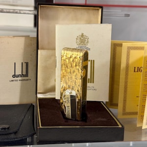 May include: A gold-coloured Dunhill lighter with a textured finish, displayed in a black box with a brown lining. The lighter is presented with paperwork and a black leather case. The Dunhill logo is visible on the paperwork.