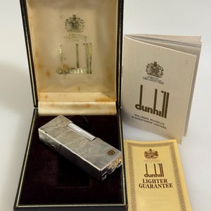 May include: A silver-toned metal Dunhill lighter with a textured finish, presented in a dark brown hinged case with a cream interior. Included are a white instruction booklet and a yellow guarantee card, both featuring the Dunhill brand logo.