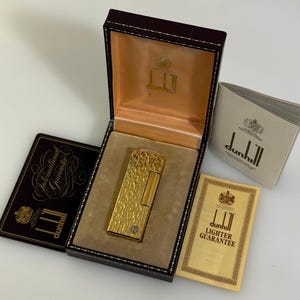 May include: A gold-toned Dunhill lighter with a textured surface, presented in a dark brown hinged box with a beige interior. Included are a black International Guarantee card, a cream-coloured Lighter Guarantee card, and a white Dunhill booklet.