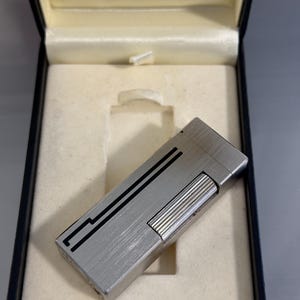 May include: A silver lighter with black detailing, displayed in a black box with a cream lining. The lighter has a rectangular shape, with parallel black lines and a textured panel.