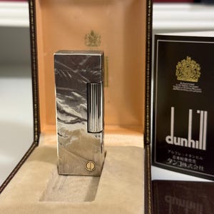 May include: A silver-toned rectangular metal lighter with a textured finish and a vertical ribbed ignition roller. The lighter features a small circular gold-coloured emblem at the base and is presented inside a brown hinged display case with tan lining.
