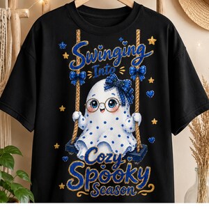 Może przedstawiać: Czarny t-shirt z grafiką białego duszka w niebieskie kropki, noszącego okrągłe okulary i niebieską kokardę we wzory, siedzącego na huśtawce linowej. Napis głosi "Swinging Into Cozy Spooky Season" niebieskimi i złotymi literami.