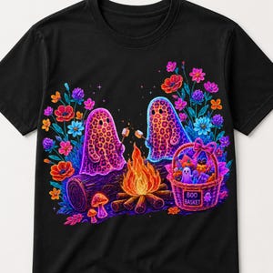 May include: A black t-shirt featuring a vibrant graphic of two leopard-print ghosts roasting marshmallows over a campfire. The scene includes colourful flowers, mushrooms, and a purple basket labelled BOO BASKET filled with various treats and items.