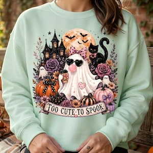 May include: A light mint green sweatshirt featuring a graphic print of a ghost wearing heart-shaped sunglasses and a polka-dot bow, surrounded by a haunted house, black cat, pumpkins, and flowers, with the text "TOO CUTE TO SPOOK" on a banner.