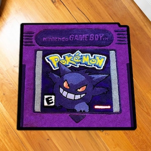May include: A purple rug shaped like a Nintendo Game Boy cartridge, featuring the Pok&eacute;mon logo and a Gengar character. The rug has a black border and is placed on a wooden floor.