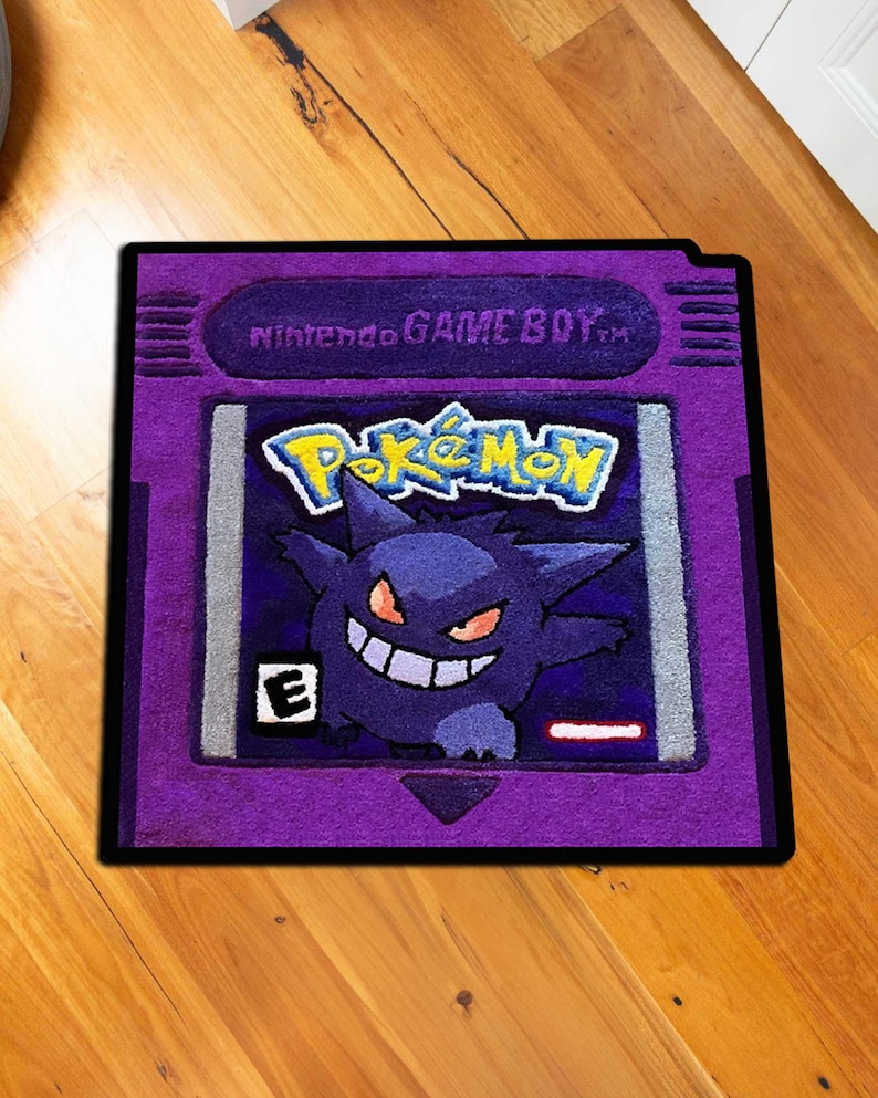 May include: A purple rug shaped like a Nintendo Game Boy cartridge, featuring the Pok&eacute;mon logo and a Gengar character. The rug has a black border and is placed on a wooden floor.