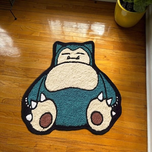 Op de afbeelding: Een teal en crèmekleurig tapijt in de vorm van de Pokémon Snorlax. Het tapijt heeft een zwarte omtrek en details, waaronder Snorlaxs gezicht, buik en voeten. Het tapijt ligt op een houten vloer.