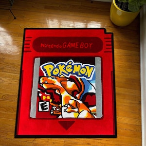 May include: A red rug in the shape of a Nintendo Game Boy cartridge. The rug displays the text "Nintendo GAME BOY" and a colourful Pokémon design. The Pokémon design features an orange and yellow creature with blue and white accents.