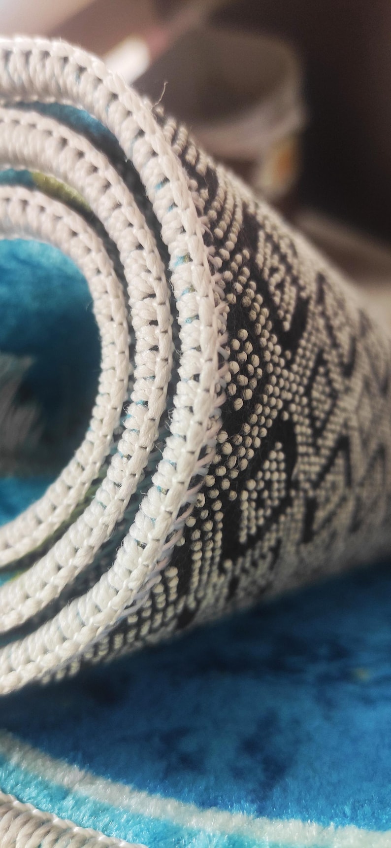 May include: Close-up of a rolled-up rug with a white, textured border. The rug features a blue and white pattern with a black and white geometric design. The rug is made of a soft, plush material.