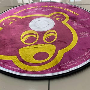 May include: Round rug featuring a stylized bear face in yellow on a maroon background. The rug is designed to resemble a CD, with text around the edges, including "Kanye West - The College Dropout."