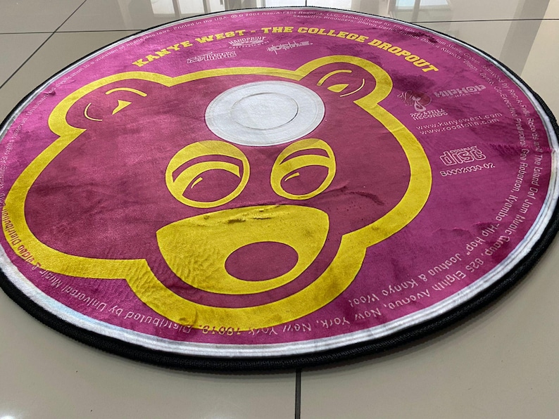 May include: Round rug featuring a stylized bear face in yellow on a maroon background. The rug is designed to resemble a CD, with text around the edges, including "Kanye West - The College Dropout."