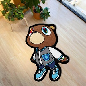 May include: A brown bear-shaped rug with a black outline. The bear wears a gray and blue letterman jacket with a blue "U" on the chest, blue jeans, and blue sneakers. The bear has large, colorful eyes and a tan muzzle.