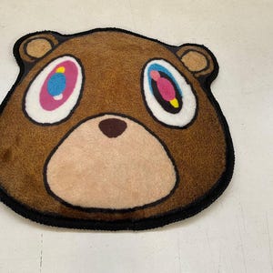 May include: A brown bear-shaped rug with a black border. The bear has large, colorful eyes with pink, blue, and yellow accents. The rug is made of a soft, plush material and is designed to resemble a cartoon bear face.