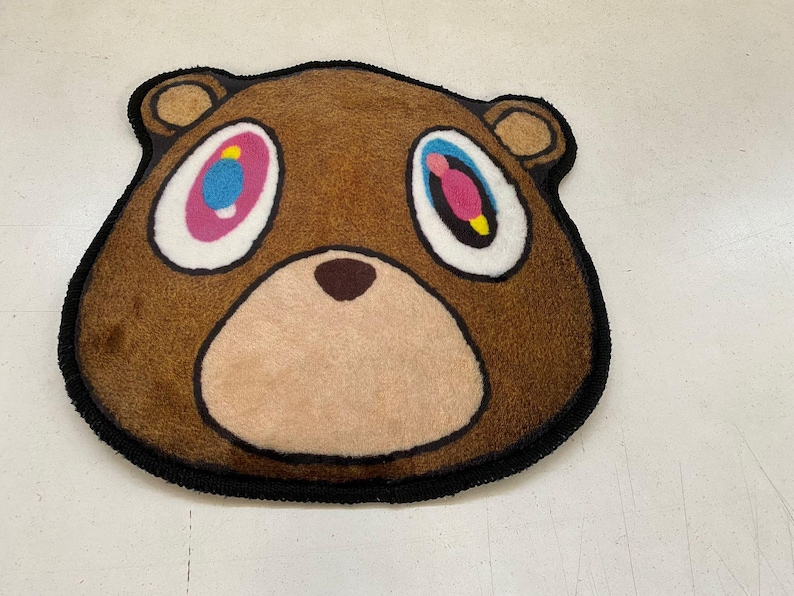 May include: A brown bear-shaped rug with a black border. The bear has large, colorful eyes with pink, blue, and yellow accents. The rug is made of a soft, plush material and is designed to resemble a cartoon bear face.